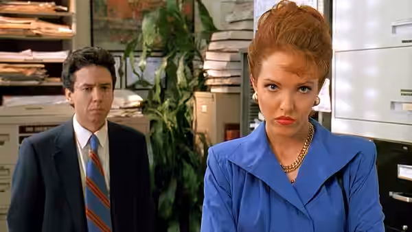 Flo (Amy Yasbeck) stands in an office as a suited man watches behind her