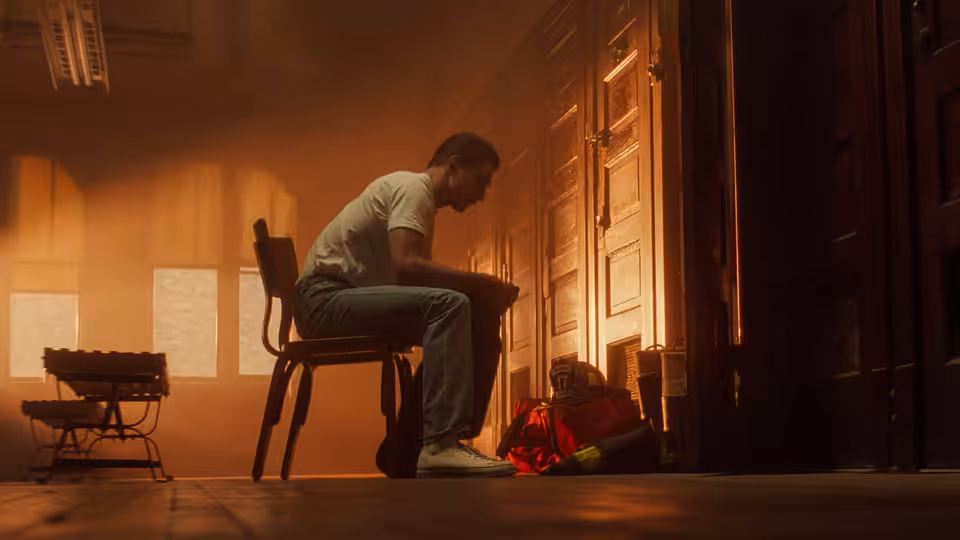 A man sits hunched in a chair in a smoke-filled room, facing a doorway