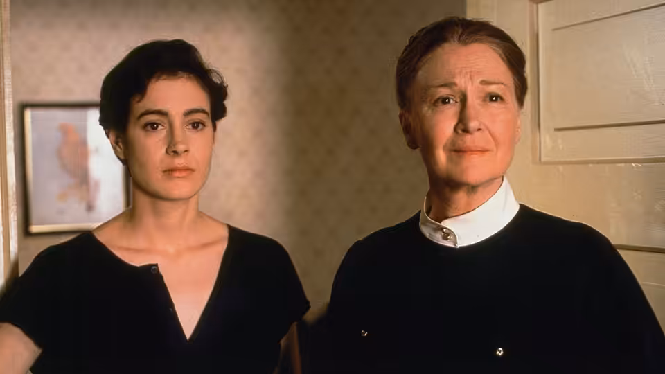 A young woman and an older woman stand together indoors, looking ahead
