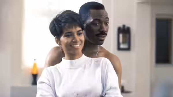 Angela smiles in front of Marcus, who stands shirtless behind her