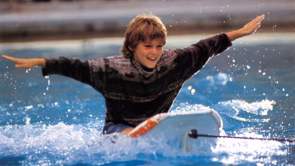 Jesse (Jason James Richter) balances on a surfboard in the water, arms spread wide