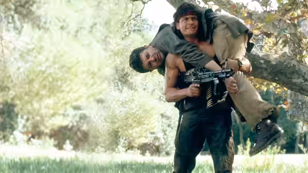 Topper Harley (Charlie Sheen) carries an unconscious man while holding a rifle in a wooded clearing