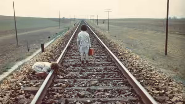 a woman walks along empty railway tracks carrying a red suitcase
