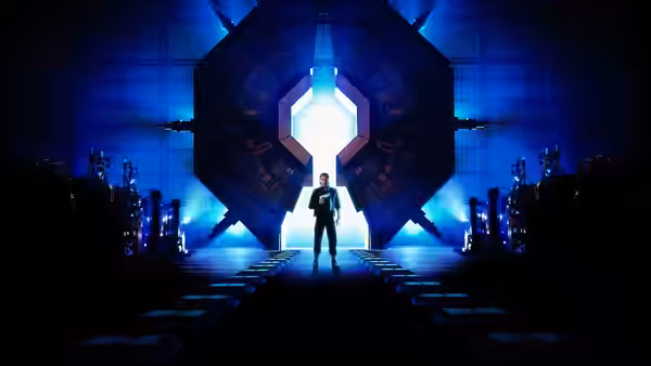 A man stands before a glowing doorway inside a vast blue-lit chamber