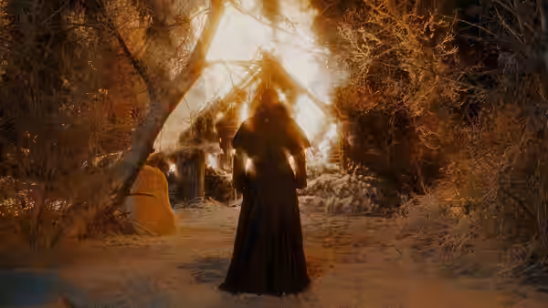 A cloaked figure approaches a burning cottage through snow-covered trees