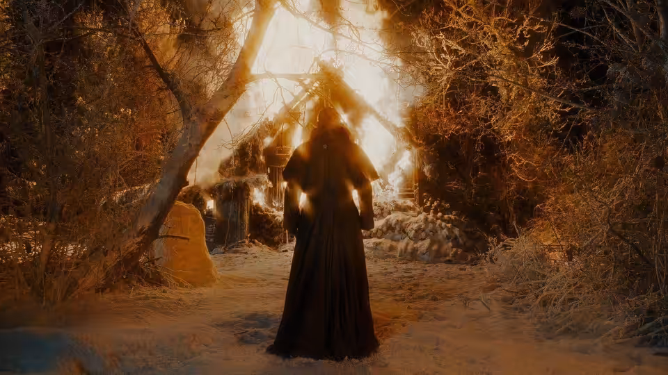 A cloaked figure approaches a burning cottage through snow-covered trees