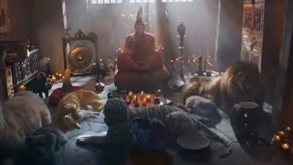 A robed figure sits among candles and resting wild animals in a hut