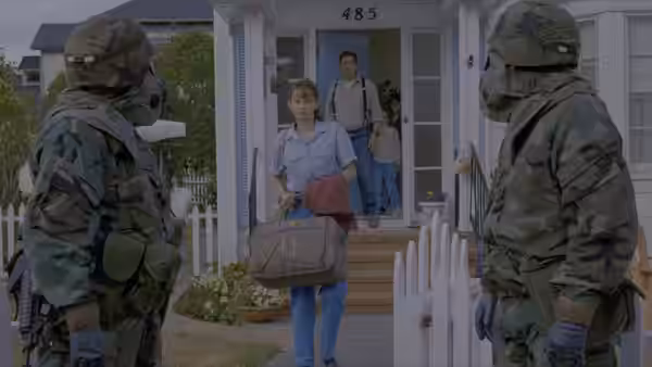 A woman carrying a bag leaves a house as soldiers stand by the gate
