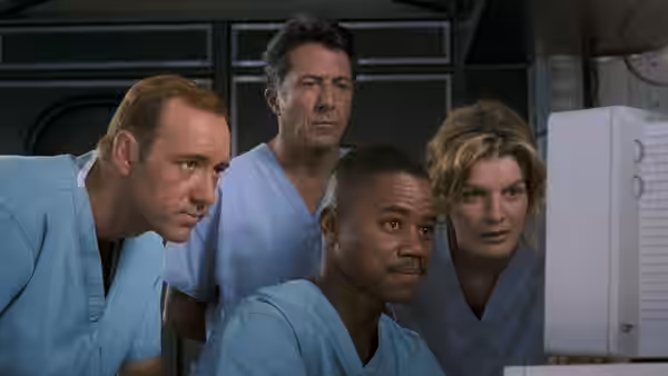 Four medical staff in scrubs watch a computer monitor