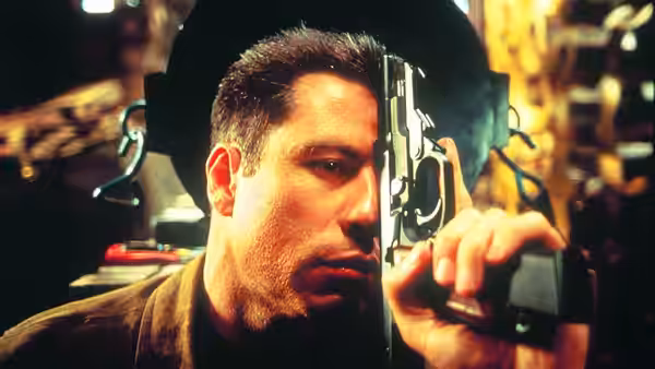 Vic Deakins (John Travolta) peers through a rifle scope in a dimly lit room