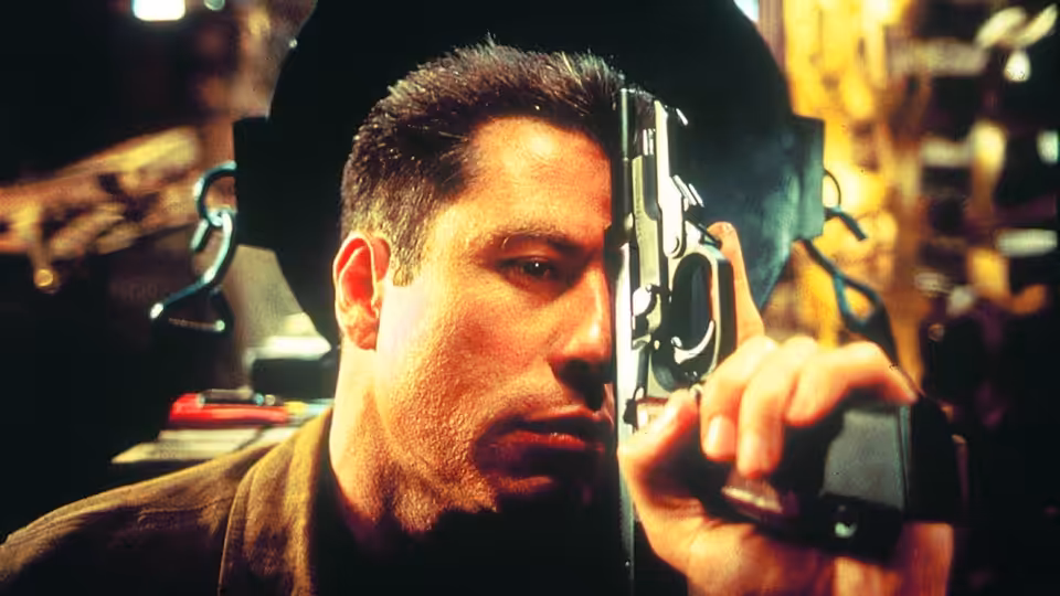 Vic Deakins (John Travolta) peers through a rifle scope in a dimly lit room