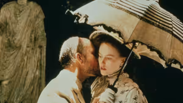A man leans close to a woman beneath a parasol beside a stone statue