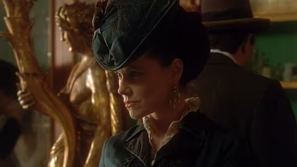 A woman in a dark hat stands indoors as a man faces away behind her