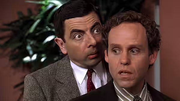 Mr. Bean (Rowan Atkinson) leans close behind a man indoors