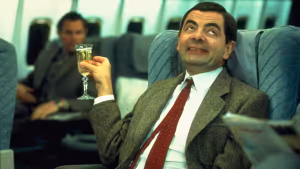 Mr. Bean (Rowan Atkinson) raises a glass while seated aboard an airplane