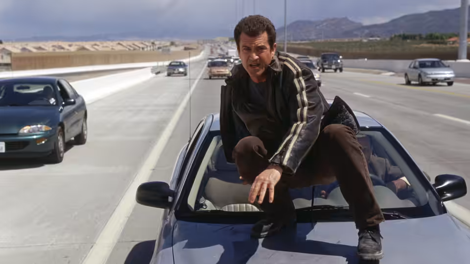 Martin Riggs (Mel Gibson) crouches atop a moving car on a busy highway