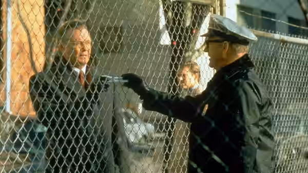 A uniformed guard points at a suited man through a chain-link fence