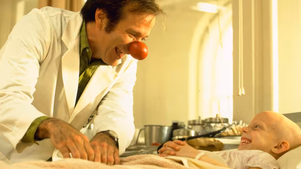 Patch Adams (Robin Williams) wears a clown nose and leans over a smiling child in a hospital bed