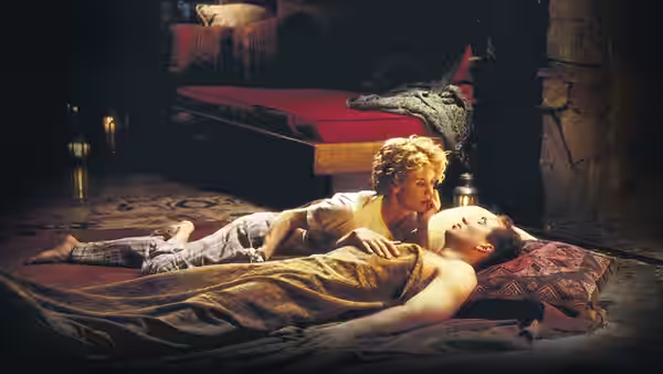A woman leans over a shirtless man lying on rugs in a dim room