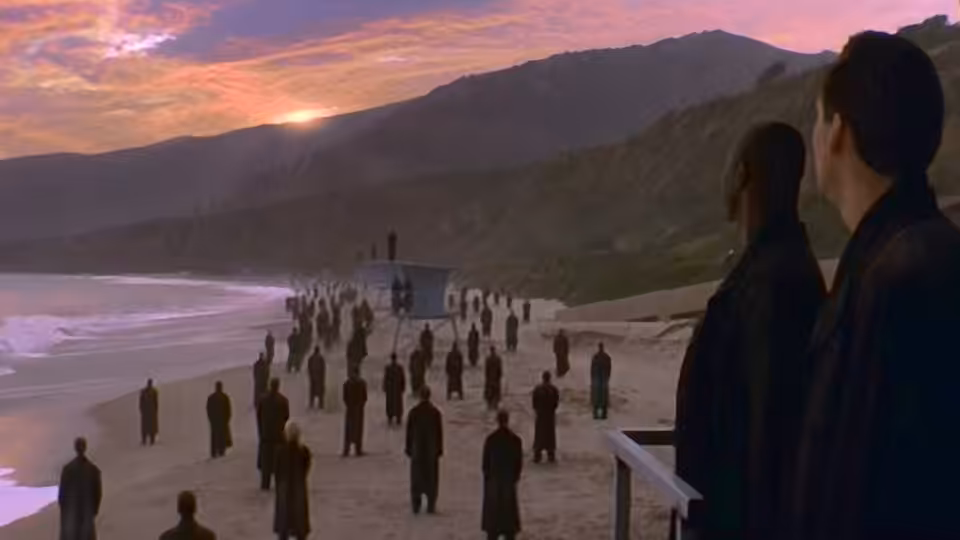 Two dark-clad figures watch a crowd gather along the beach at sunset
