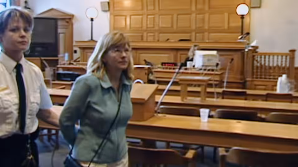 A woman stands beside a uniformed officer inside a wooden courtroom