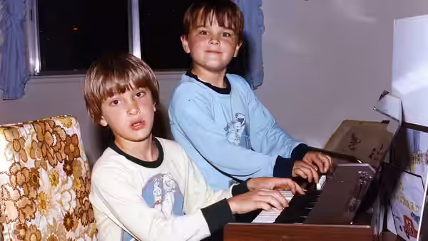 Two boys sit at an electronic keyboard in a living room