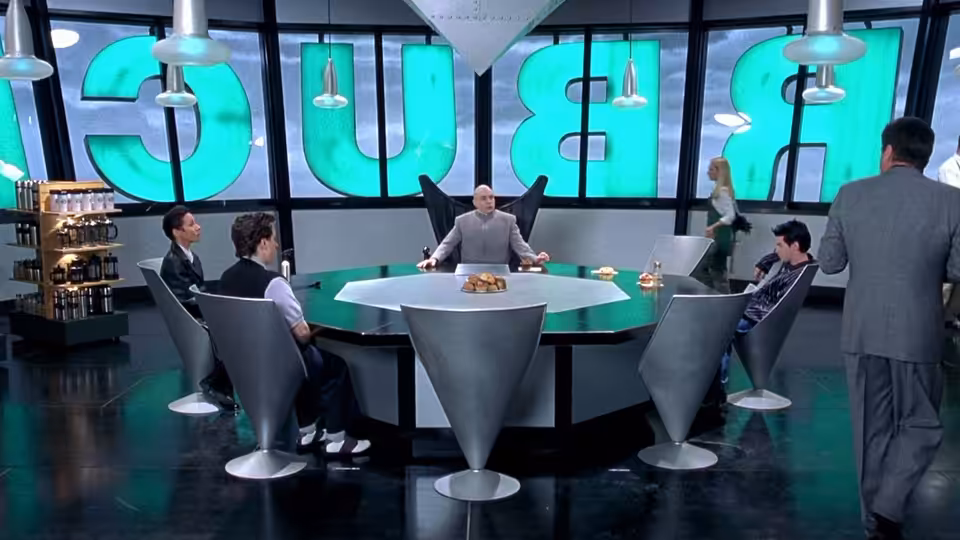 Dr. Evil addresses his associates around a table in the BU headquarters