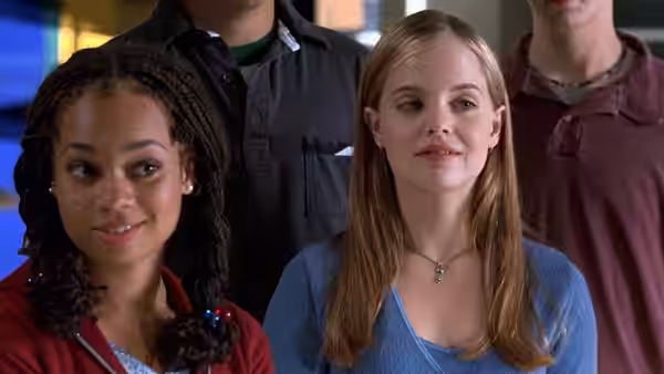 Two young women stand together indoors with other students behind them