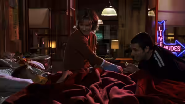 Layla sits on a bed with Sonny as Julian lies beneath the blankets