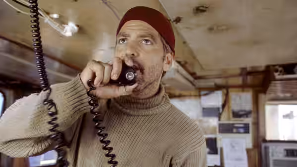 A bearded fisherman in a red cap speaks into a radio inside the boat