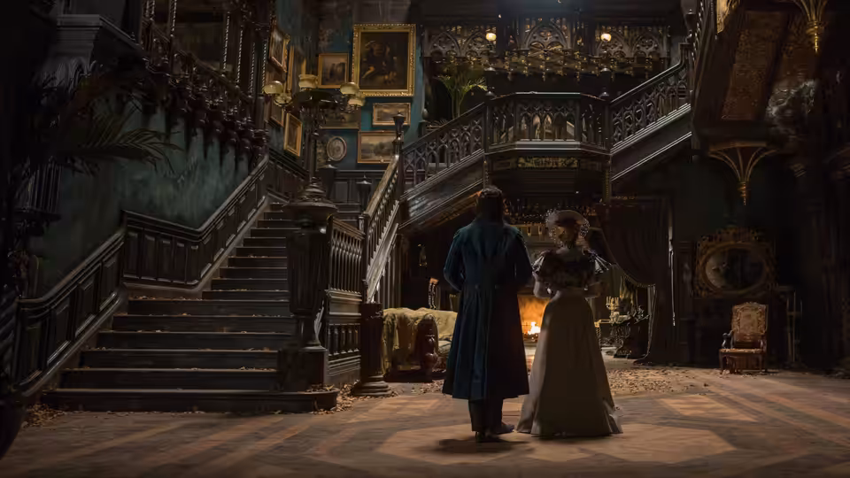 A man and woman stand beneath the sweeping staircase in a dim, ornate mansion