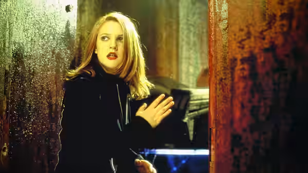 A blonde woman in black peers around a rusted doorway in a dim industrial room