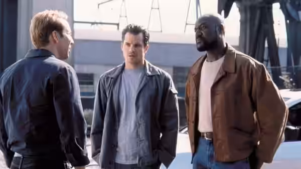 Three men talk outside an industrial building beside parked vehicles