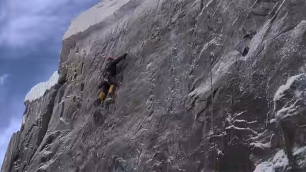 A climber in yellow trousers scales a sheer ice-covered mountain face on a rope