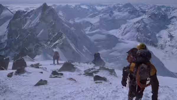 A climber with a loaded pack stands above a snowy mountain camp as others cross the ridge