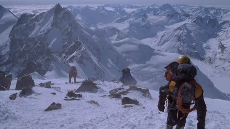 A climber with a loaded pack stands above a snowy mountain camp as others cross the ridge