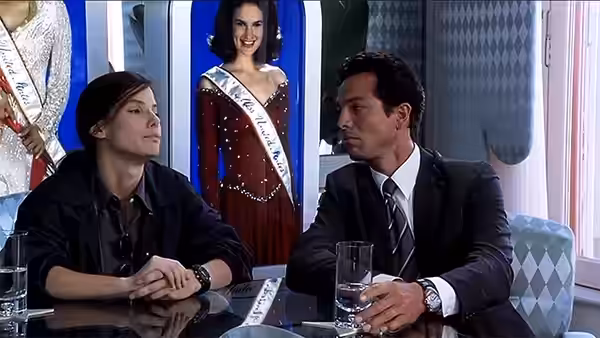 Two men sit at a table beside a smiling pageant contestant in a sash