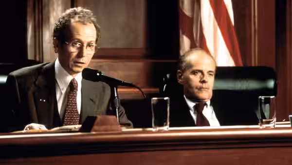 A bespectacled committee member speaks into a microphone beside a seated colleague