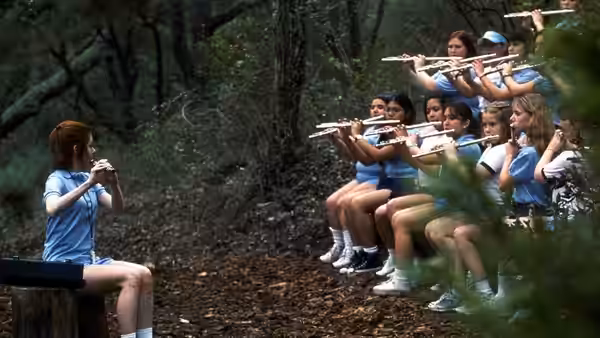 A seated flute player faces a row of uniformed girls playing flutes in the woods