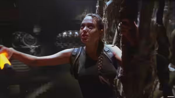 Lara Croft (Angelina Jolie) reaches across a dim chamber beside an ancient stone structure