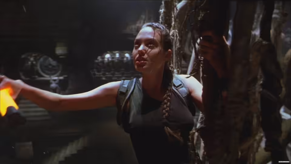 Lara Croft (Angelina Jolie) reaches across a dim chamber beside an ancient stone structure