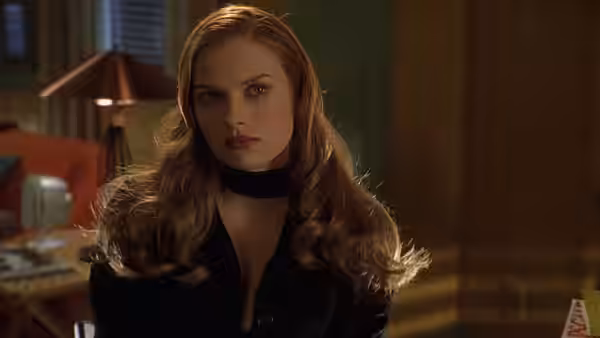 A blonde woman in a black choker sits in a dimly lit room