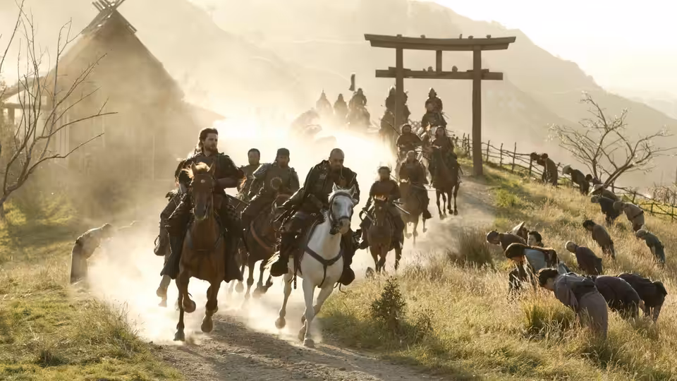 Samurai riders charge down a dusty village road beneath a wooden gate