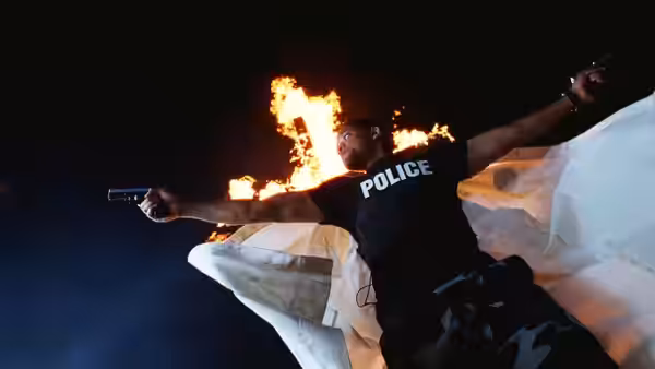 A police officer falls through the air, aiming two pistols with flames behind him