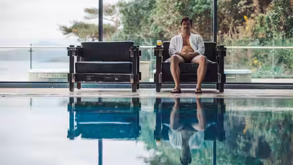 man sitting shirtless on a chair beside an indoor swimming pool