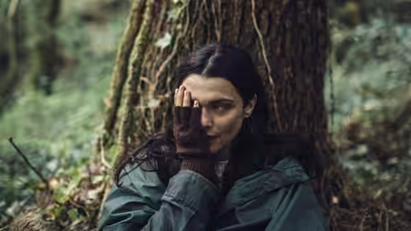 woman in a green coat resting against a tree in the woods