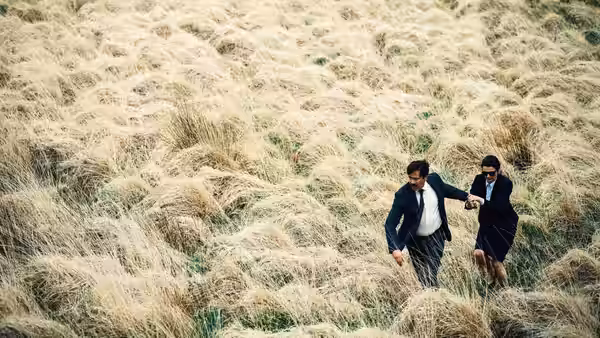 man and woman in suits running through a field of tall dry grass