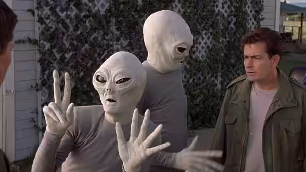 Two grey aliens gesture beside a concerned man in a backyard