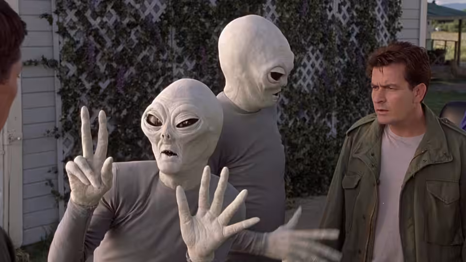 Two grey aliens gesture beside a concerned man in a backyard