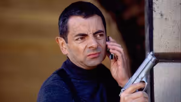Johnny English (Rowan Atkinson) speaks on a phone while holding a pistol beside a doorway